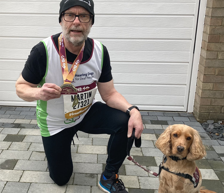 A man in in running clothing kneeling while holding a medal. A dog is sitting beside him.