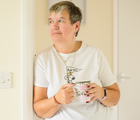 A woman is leaning against a wall with a cup of hot drink in her hands