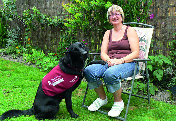 Jackie is sitting on a chair in the garden with black Labrador hearing dog Quattro sitting to the side of her