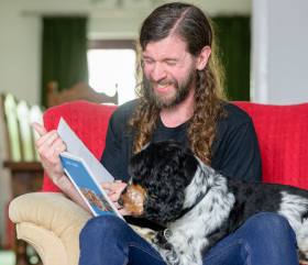 A smiling man and cocker spaniel dog sitting ona  sofa looking at a book