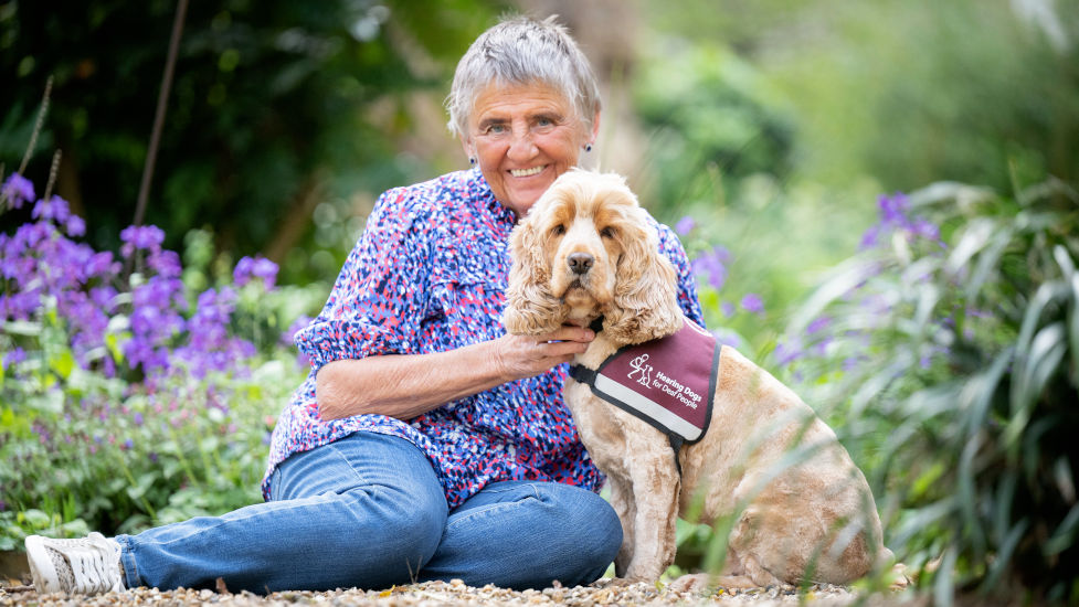 A woman sitting on the ground beside a light coloured dog which is wearing a burgundy hearing dogs jacket. They are surrounded by greenery and purple flowers