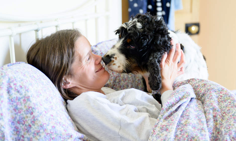 A smiling woman lying on a bed being after been awoken by a hearind dog