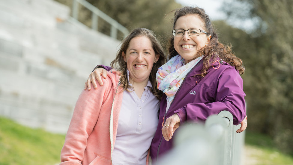 A woman (Lucy) wearing a purple jacket and glasses is standing with her right arm around her daughter's shoulder (Pip). Both women are smiling.