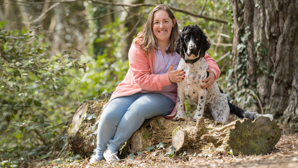 A young woman (Pip) is sitting on a large log with her arms around a black and white dog (Gino). They are in a woodland area.