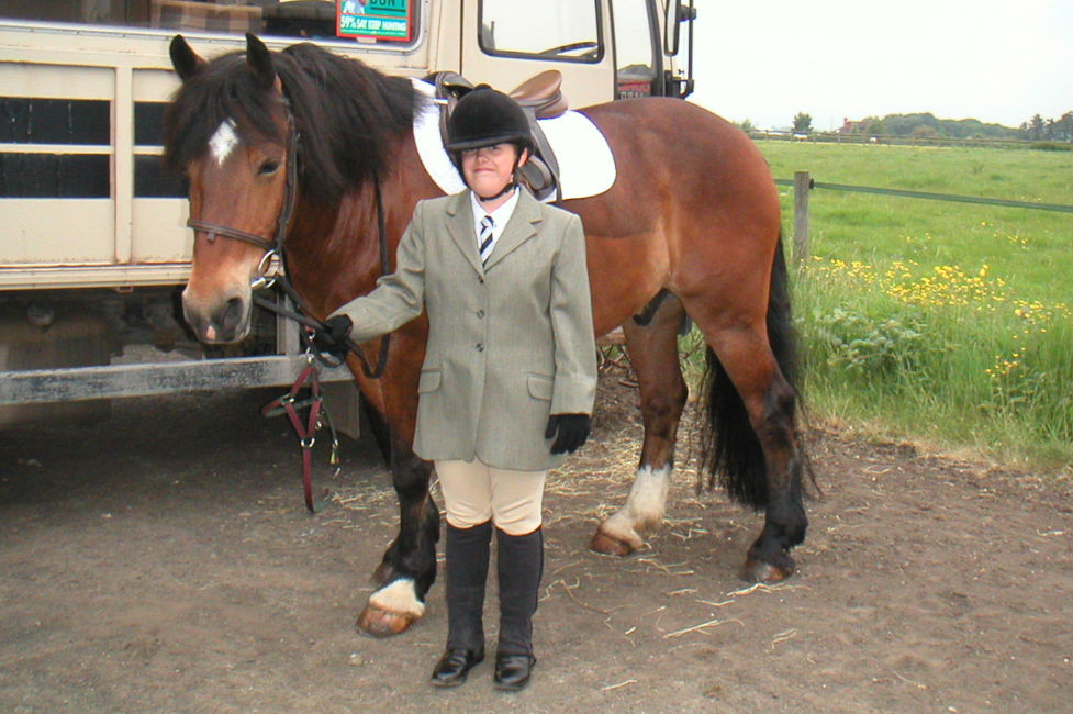 A young girl (Pip) standing with a brown horse holding the reins. A green field with yellow flowers is in the background.