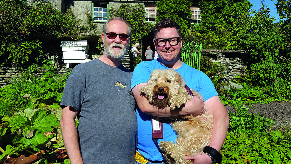 Phil and Lee standing side by side on a summer's day with Lee holding hearing dog Juno who is wearing her burgundy jacket
