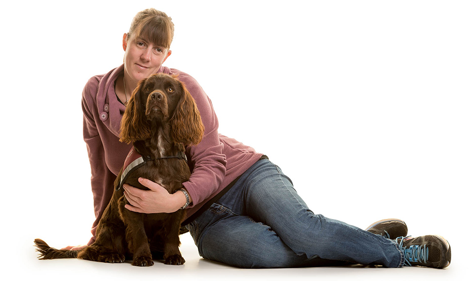 Jen sitting on the floor, with her arm around cocker spaniel hearing dog Dram, who is sitting in front of her