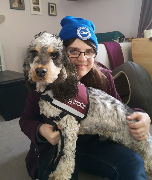 Hearing dog Archer in his burgundy jacket is sitting on Rachel's lap