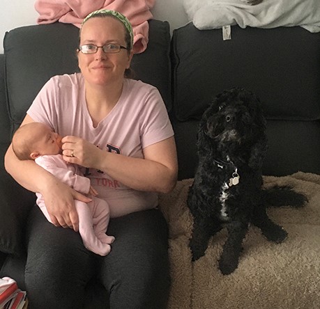 Kirsten is sitting on a sofa and holding baby Noelle in her arms with hearing dog Pickle sat beside her