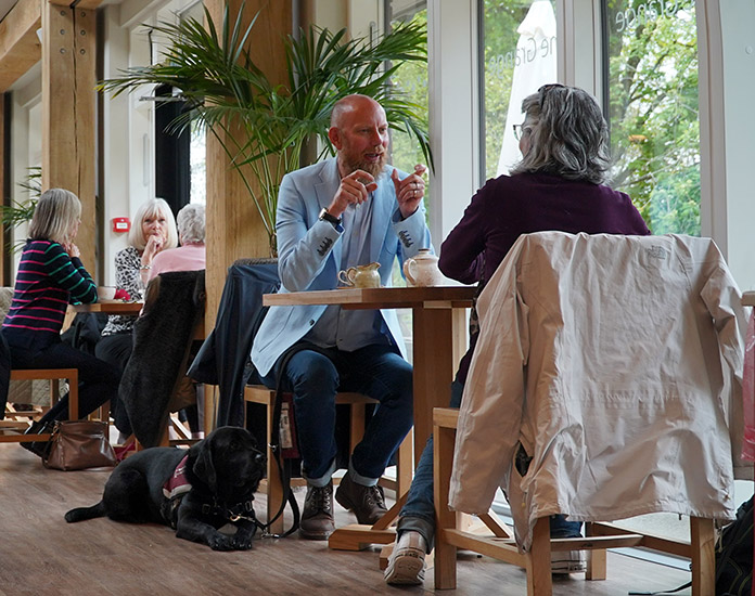 Ian is sitting at a table in The Grange Restaurant talking with hearing dog Vectra lying by his feet.