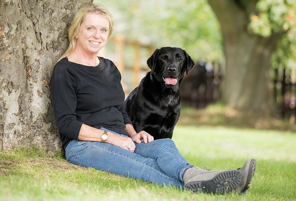 Carolann is sitting on the grass next to a tree with hearing dog Walnut next to her