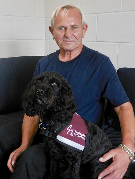 David and hearing dog Asti (a black cockapoo) are sitting on a sofa together. David's arm is around Asti.
