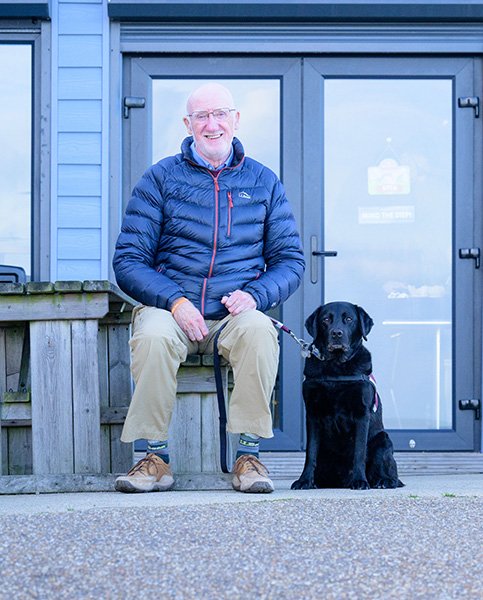 Deryck is sitting on a bench outside at a cafe and is looking towards the camera, smiling. Hearing dog Elroy is sitting on the floor to Deryck's left 