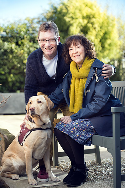 David and Linda are sitting next to each other on a bench with hearing dog Fidget, a yellow Labrador, sitting on the floor in front of them.