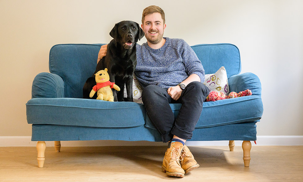 Ralph and black Labrador Winnie are sitting on a sofa. Next to Winnie is a cuddly toy Winnie the Pooh.